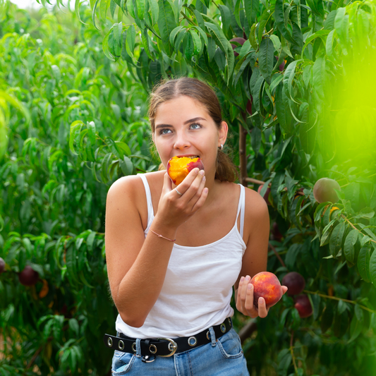 Tan girl in white tank top eating a peach in an orchard